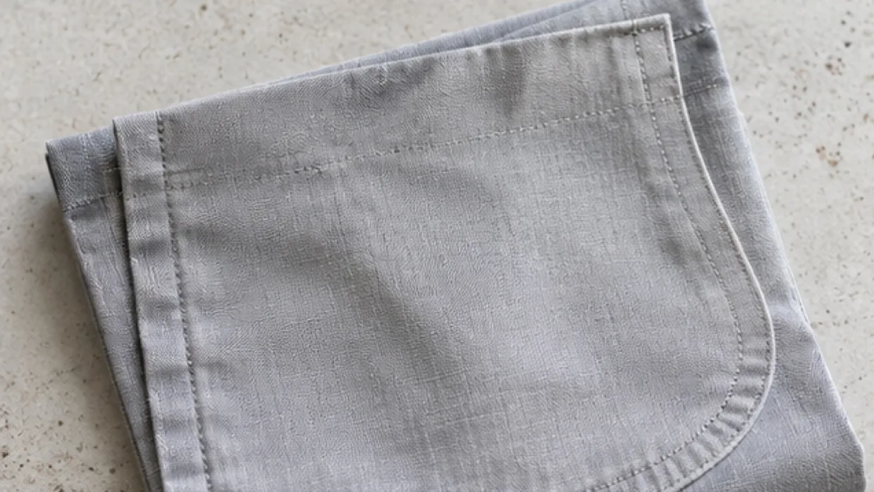 A folded grey pocket bag cloth with curved stitched edge on a pale stone surface