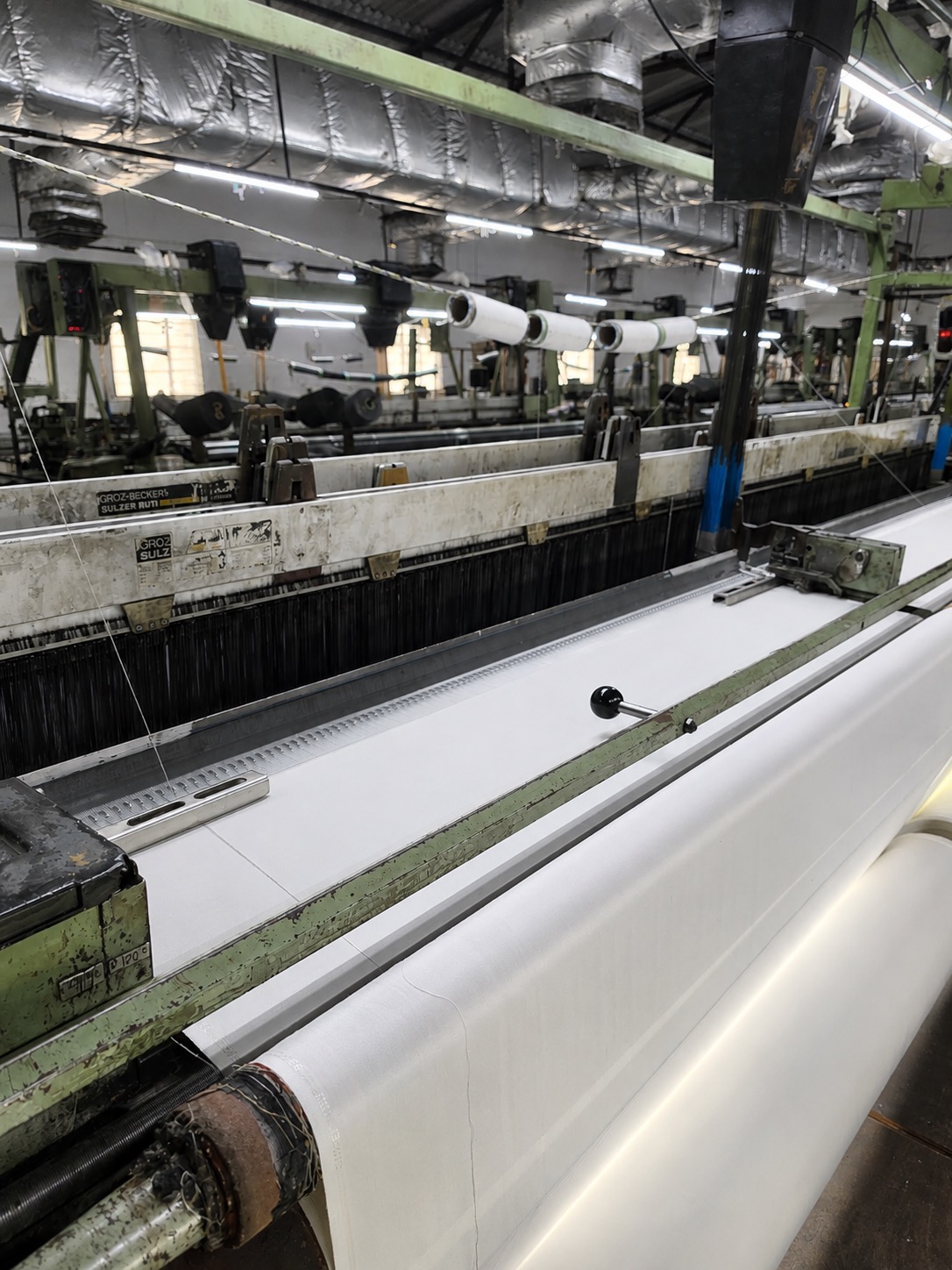 Long corridor between rows of looms in a weaving hall, with overhead ducting running the length of the aisle