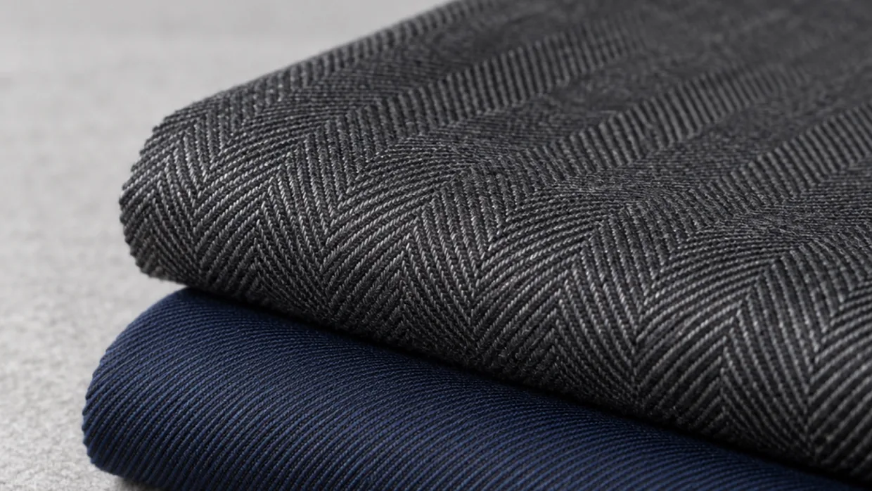 A charcoal herringbone swatch folded on top of a navy twill swatch on grey flannel